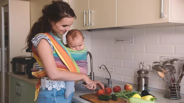 Maminka krájí zeleninu s dítětem v nosičce na kuchyňské desce.