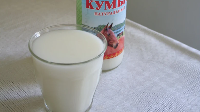 Sklenička kumysu vedle lahve s nápisem Kumys na bílém ubrusu.