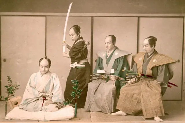 Japonská scéna seppuku s samurajem držícím meč nad mužem.