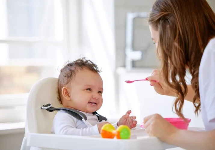 Maminka krmití šťastné dítě v jídelní židli.