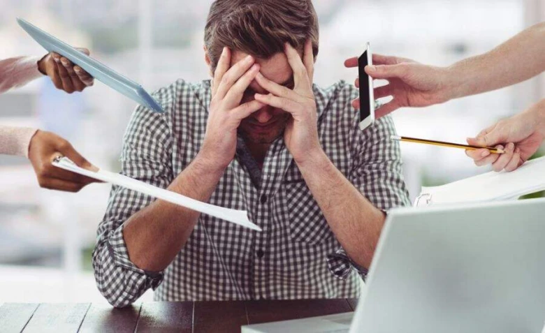 Muž v kanceláři přetěžovaný prací s dokumenty a zařízeními.