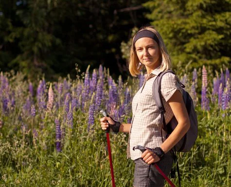 Žena s turistickými holemi stojí mezi fialovými květinami v přírodě.