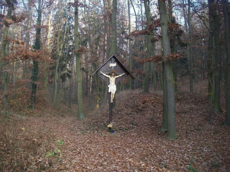 Kříž s Ježíšem na křižovatce v lese pod šedou oblohou.