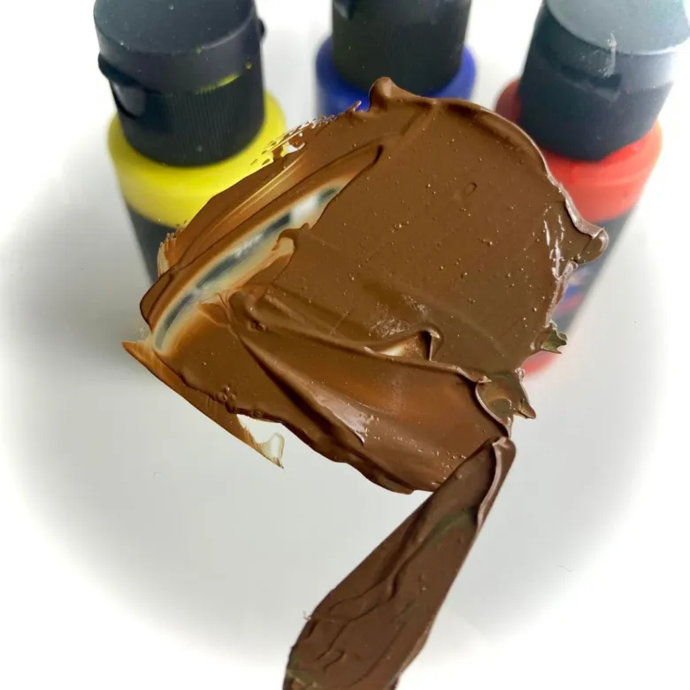 Hnědá barva namíchaná na lžíci s barevnými lahvičkami v pozadí.