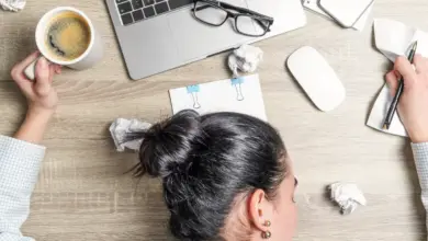 Žena sedí u stolu s laptopem a kávou v ruce.