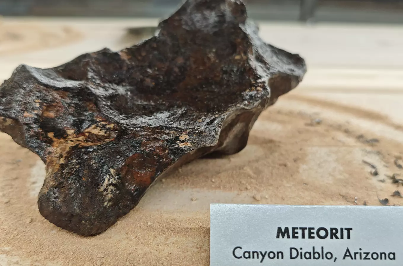Meteorit z Arizony na pískovém podkladu s popisek.