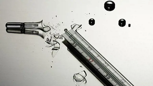 Rozbitý skleněný teploměr s rozlitým rtutí na bílém povrchu.