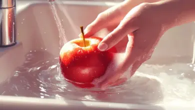 Ruka umývá červené jablko pod tekoucí vodou v dřezu.