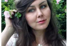 Žena drží růži s pěti lístky u tváře na pozadí zelených listů.