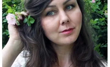Žena drží růži s pěti lístky u tváře na pozadí zelených listů.