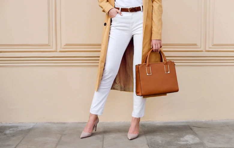Žena v elegantním neformálním oblečení s taškou stojí u zdi.
