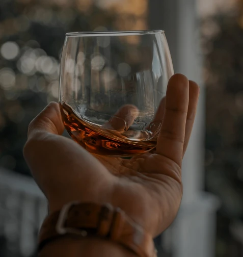 Ruka drží sklenici s hnědým nápojem, pravděpodobně koňak nebo whisky.