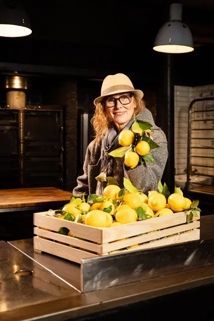 Žena drží větve s citrony nad krabicí čerstvých citrusů.