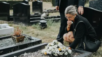 Žena v černém pláštíku smutečně klečí u hrobu s květinami.