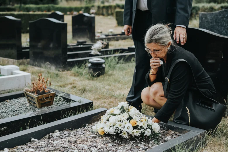 Žena v černém pláštíku smutečně klečí u hrobu s květinami.