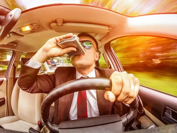 Muž pije alkohol za volantem auta v pohybu.