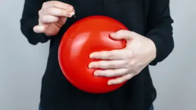 Člověk drží červenou nafukovací kouli, kterou bodne jehlou.