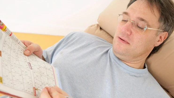 Muž řeší sudoku v časopisu, zatímco leží na gauči.