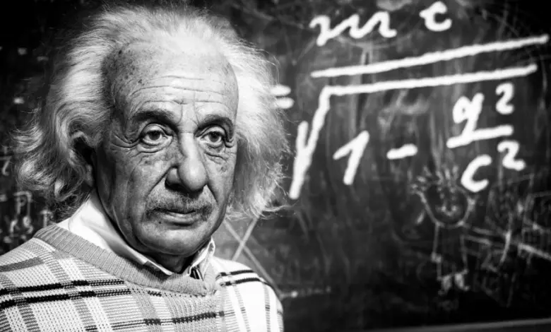 Portrét Alberta Einsteina před tabulí s fyzikálními rovnicemi.