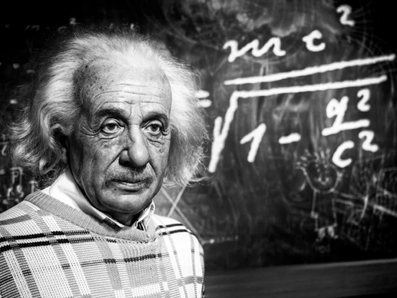 Portrét Alberta Einsteina před tabulí s fyzikálními rovnicemi.