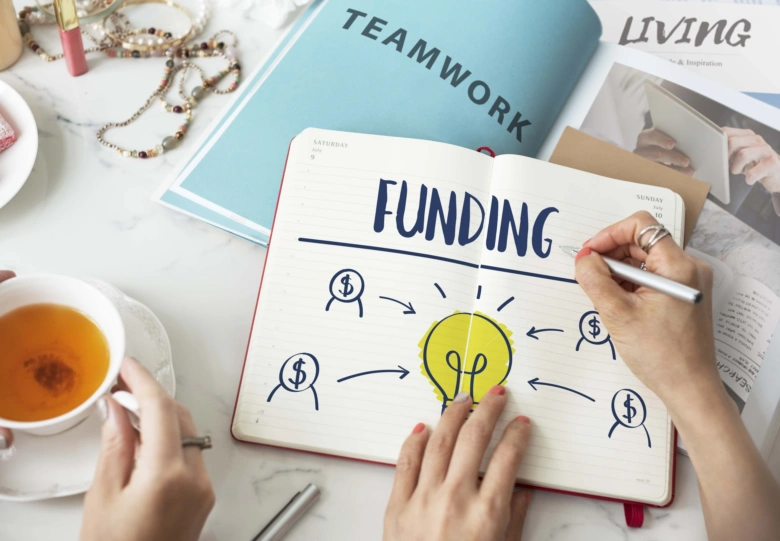 Ruce píšou slovo FUNDING v poznámkovém sešitu s ikonami peněz.