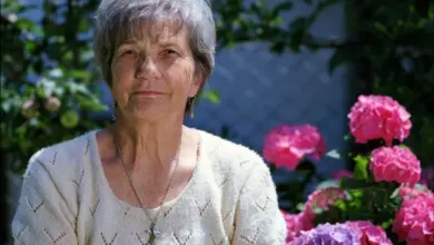 Starší žena stojí venku mezi kvetoucími růžemi.