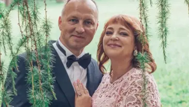 Manželé v formálním oblečení stojí venku mezi zelenými větvemi.