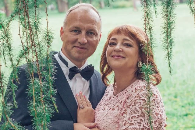 Manželé v formálním oblečení stojí venku mezi zelenými větvemi.