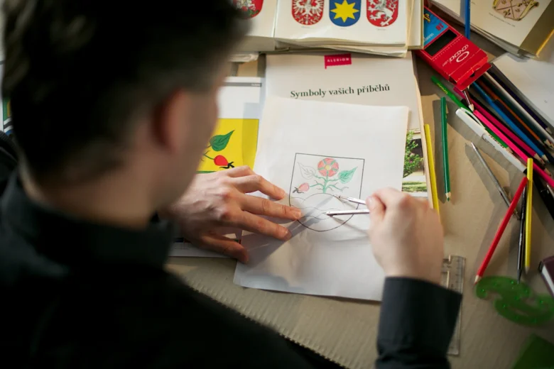 Člověk kreslí erb s květinou na papíru mezi pomůckami.