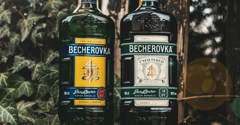 Dvě lahve Becherovky před zelenou stěnou s liánami.