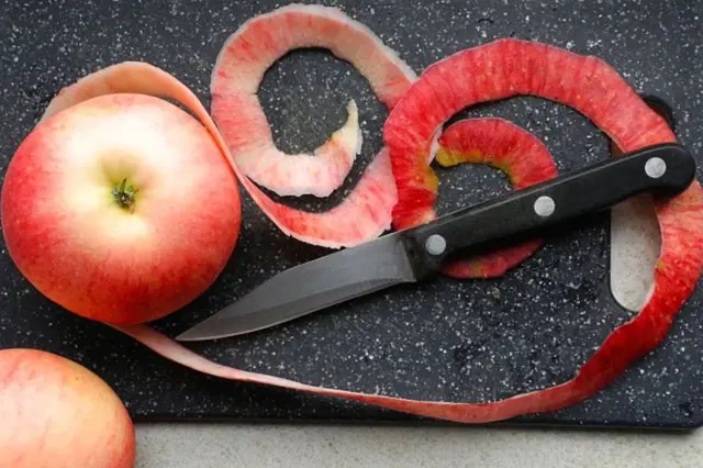 Červené jablko s odloupenou kůrou a nožem na tmavém prkénku.