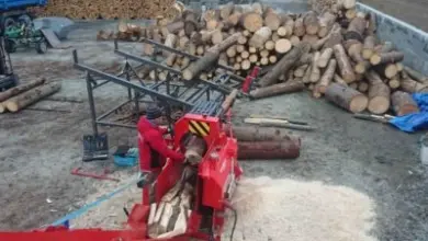 Červený stroj na rozdělování dřeva s hromadami kmenů v pilníku.