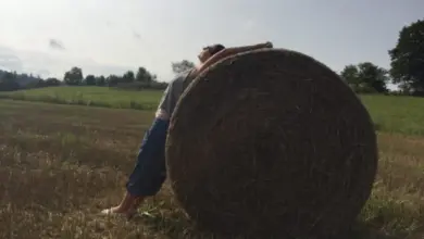 Osoba se opírá o slámovou balu na louce.