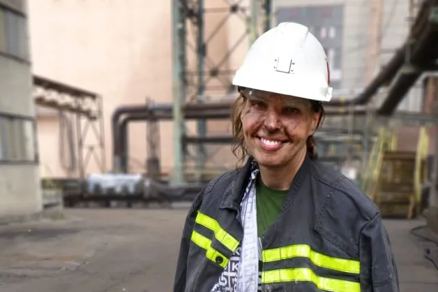 Usměvavá žena v průmyslovém prostředí s ochrannou helmkou.