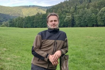 Muž stojí na louce s lesy v pozadí.