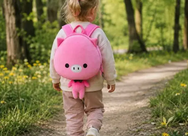 Dívka s růžovým batohem v tvare prasátka chodí lesní cestou.