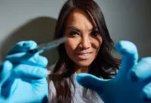 Žena v laboratorním plášti s modrými rukavicemi drží chirurgický nástroj.
