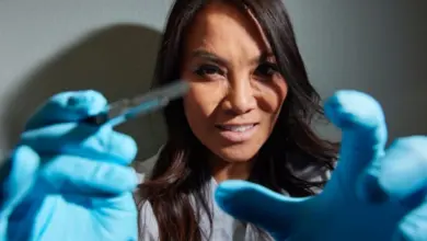 Žena v laboratorním plášti s modrými rukavicemi drží chirurgický nástroj.