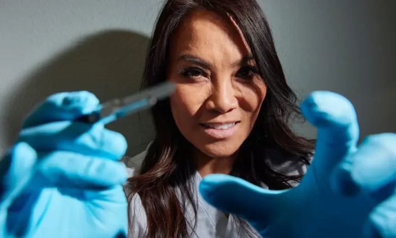 Žena v laboratorním plášti s modrými rukavicemi drží chirurgický nástroj.