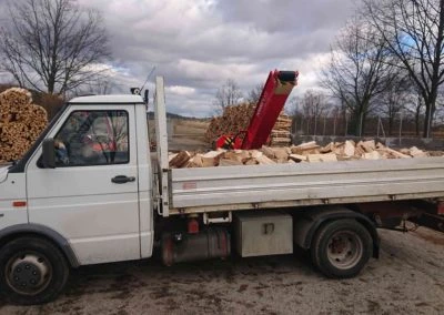 Bílý nákladní vůz s dřevem a hydraulickým nakladačem venku.